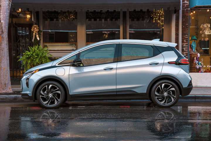 Static side profile shot of a silver 2022 Chevrolet Bolt EV parkerd outside of some shops in a city.