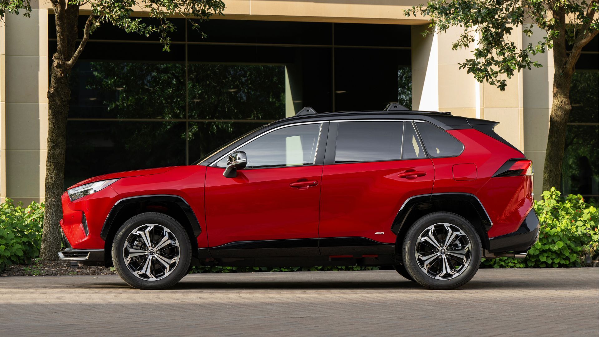 Static side profile shot of a red 2025 Toyota RAV4 PHEV XSE parked outside of a modern building.